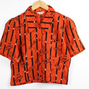 May include: Orange short-sleeved button-down shirt with black vertical stripes and a repeating pattern. The shirt has a collared neckline and is hanging on a wooden hanger.