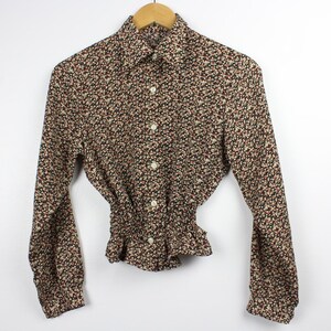 Puede incluir: Blusa de manga larga con estampado floral en marrón, negro y rojo. La blusa tiene cuello, cierre de botones y cintura fruncida. Cuelga de una percha de madera sobre un fondo blanco.
