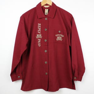 May include: A long-sleeved, red button-down shirt with embroidered text that reads "REPLY PAID" on the left side and "REPLY PAID" with a crown design on the right side.