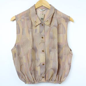 May include: A sleeveless, button-up blouse with a geometric pattern in shades of pink, beige, and brown. The blouse has a collar with a lace trim and a gathered hem.