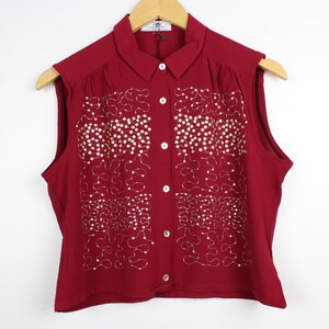May include: A sleeveless burgundy button-down shirt featuring a collar and a decorative pattern of silver-toned sequins and metallic thread embroidery on the front panels. The garment is displayed on a wooden hanger against a plain white background.