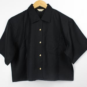 May include: A black short-sleeved button-down shirt with gold buttons. The shirt has a pocket on the left side.