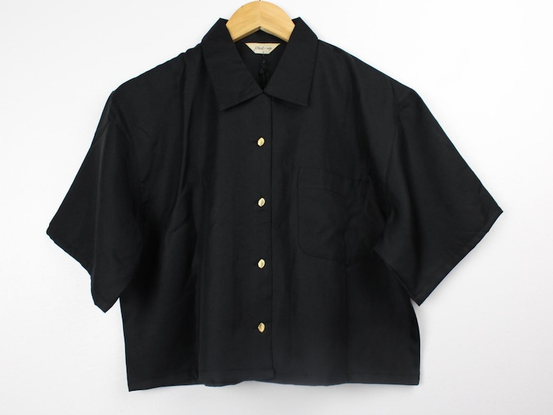 May include: A black short-sleeved button-down shirt with gold buttons. The shirt has a pocket on the left side.
