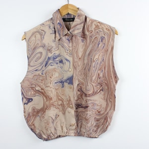 May include: Sleeveless button-up shirt with a swirling pattern in shades of beige, brown, and blue. The shirt has a collared neckline and is hanging on a wooden hanger. The bottom of the shirt is gathered.