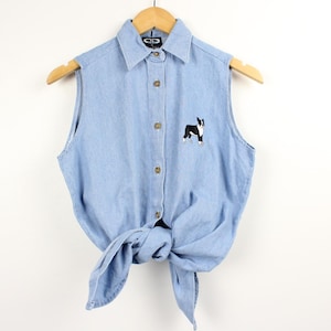 Op de afbeelding: Lichtblauwe mouwloze spijkerblouse met een knoopsluiting aan de voorkant en een knoop aan de taille. De blouse heeft een kleine geborduurde zwart-witte hond op de borst. De blouse hangt aan een houten hanger.