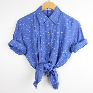 May include: A blue button-down shirt with a white repeating pattern of small shapes. The shirt features a pointed collar, a front button placket, and long sleeves that are rolled up. The bottom hem is tied in a knot at the center front.