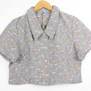 May include: A light gray, short-sleeved button-down shirt with a floral pattern in shades of yellow, orange, and blue. The shirt has a classic collar and is hanging on a wooden hanger.