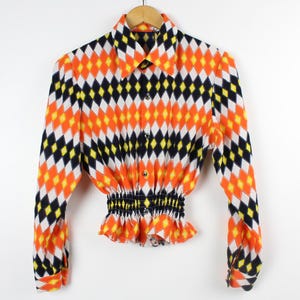 Puede incluir: Una camisa de manga larga con un patrón geométrico en naranja, negro, blanco y amarillo. La camisa tiene un cuello abotonado y una cintura ajustada.