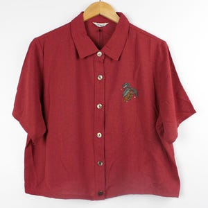 Puede incluir: Una camisa roja de manga corta con botones y un diseño bordado de dos pájaros en la parte delantera.