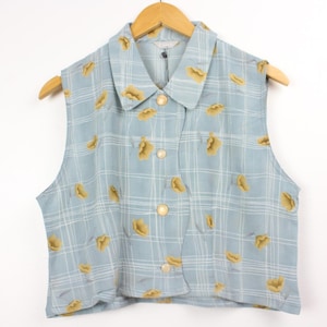 May include: A sleeveless button-down vest in a light blue color featuring a white plaid pattern and a repeating yellow floral print. The garment has a classic collar, a front button closure with round buttons, and a straight hemline.