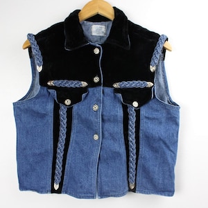 Puede incluir: Un chaleco vaquero con cuello y detalles de terciopelo negro. El chaleco presenta botones plateados y ribetes trenzados azules. El chaleco es sin mangas y cuelga de una percha de madera.