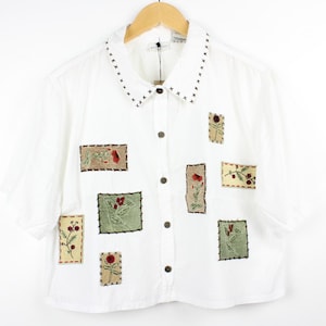 May include: A white short-sleeved button-down shirt featuring a collar with black cross-stitch detailing. The front of the shirt is decorated with several rectangular patches in shades of tan and green, each embroidered with floral motifs.