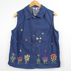 May include: A sleeveless denim shirt with embroidered flowers in pots along the bottom and small butterflies scattered across the front. The shirt has a button-down front and a collared neckline. The shirt is hanging on a wooden hanger.