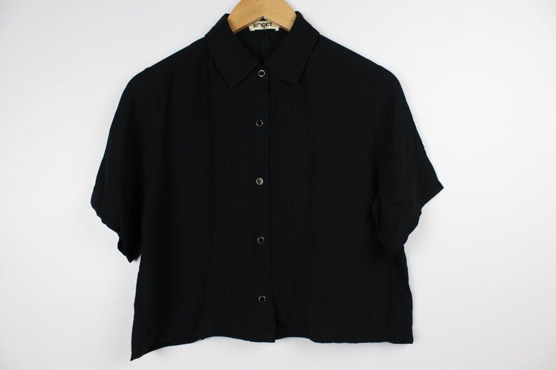 May include: A black short-sleeved button-down shirt with a collared neckline. The shirt has a slightly cropped length and a textured fabric.
