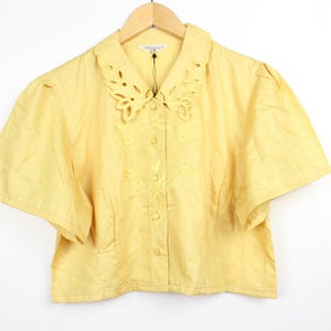 May include: A pale yellow short-sleeved blouse featuring a pointed collar with intricate cut-out embroidery details. The garment has a button-down front closure and tonal floral embroidery on the chest area, hanging on a wooden hanger.