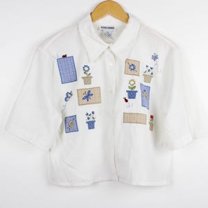 May include: A white short-sleeved button-down shirt featuring embroidered patches of blue and tan checkered squares, potted flowers, dragonflies, and ladybugs. The shirt has a classic collar and is displayed on a wooden hanger with the label Alfred Dunner.