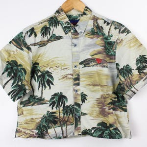 Puede incluir: Una camisa blanca de manga corta con botones y estampado tropical con palmeras y una escena de playa. La camisa tiene un corte holgado y una longitud recortada.