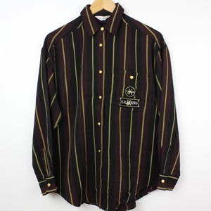 Può includere: Una camicia a maniche lunghe con bottoni e strisce verticali nere e verdi. La camicia ha una toppa sul petto sinistro che dice "K-PLINKO".