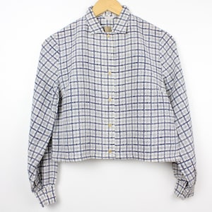 May include: A long-sleeved button-down shirt featuring a white and navy blue plaid grid pattern. The garment has a classic collar, a straight hemline, and light-colored buttons running down the center front, all displayed on a wooden hanger.