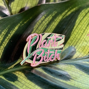 May include: A gold-colored enamel pin with a green and pink leaf design. The words "Plant Bitch" are written in pink on the leaf.