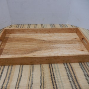 May include: A rectangular wooden serving tray with a light brown finish. The tray has cut-out handles on each side. The wood grain is visible, adding to its natural aesthetic. The tray is empty and sits on a striped surface.