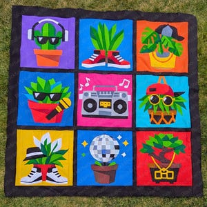 Puede incluir: Un edredón colorido con nueve paneles cuadrados que representan plantas al estilo pop art. Cada panel muestra una planta diferente, como un cactus o un bonsái, adornada con accesorios como auriculares, gafas de sol y una radio. El edredón tiene un borde negro.