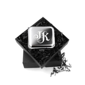 Op de afbeelding: Een zilveren compact spiegel met een gegraveerd ontwerp met de initialen "JK" in een decoratief handschrift. De spiegel zit in een zwarte cadeaudoos met zwart geribbeld papier.
