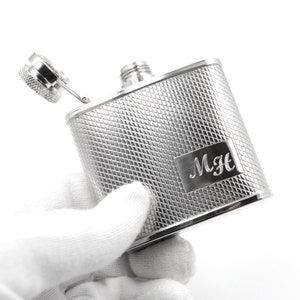 Puede incluir: Una petaca de plata con un patrón de diamante y una inscripción personalizada que dice "MH". La petaca está abierta y la tapa está quitada.