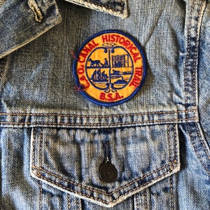 May include: A denim jacket with a circular patch. The patch is yellow with a blue border and features the text "GEO. CANAL HISTORICAL TRAIL B.S.A." along with various illustrations. The jacket has a buttoned pocket and a button closure.