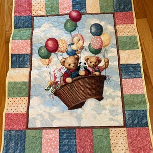 May include: A quilted baby blanket featuring a whimsical scene of teddy bears in a hot air balloon basket, floating among colorful balloons. The border has a patchwork design with squares of pink, blue, green, and cream. The background is a light blue sky with fluffy clouds.