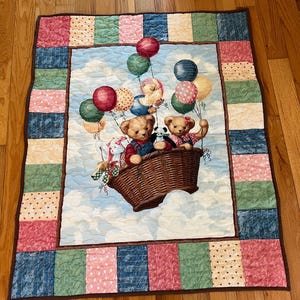 May include: A quilted baby blanket featuring a whimsical scene of teddy bears in a hot air balloon basket, floating among colorful balloons. The border is a patchwork of various patterned fabrics in shades of pink, blue, green, and beige.