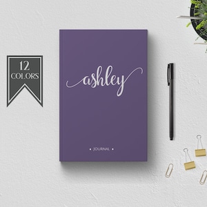Pode incluir: Um diário roxo com o nome "ashley" escrito em caligrafia branca. O diário tem um rótulo preto que diz "12 COLORS" e a palavra "JOURNAL" está impressa em branco abaixo do nome.