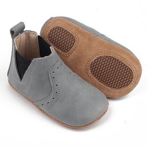 May include: Grey suede baby booties with brown suede soles. The booties have an elastic back and a decorative stitch pattern.