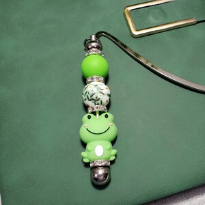 May include: A green frog-themed bookmark with a silver metal top. The bookmark features a green frog charm, green and white beads, and a silver metal bookmark. The bookmark is resting on a green notebook.