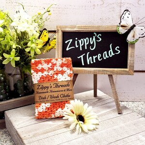 May include: An orange and white crocheted dishcloth is displayed with a small chalkboard sign that reads "Zippy's Threads". The image includes a wooden crate with flowers and a decorative butterfly.