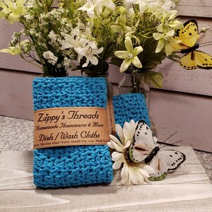 Puede incluir: Pa&ntilde;os de cocina de ganchillo turquesa con una etiqueta marr&oacute;n que dice "Zippy's Threads Homemade Housewares & More Dish/Wash Cloths". La imagen incluye flores blancas y mariposas.
