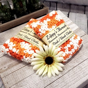 May include: Two handmade dish cloths in orange and white crochet, labeled "Zippy's Threads". The cloths are displayed with a white and yellow flower. The text on the label reads "Homemade Housewares & More Dish/Wash Cloths".