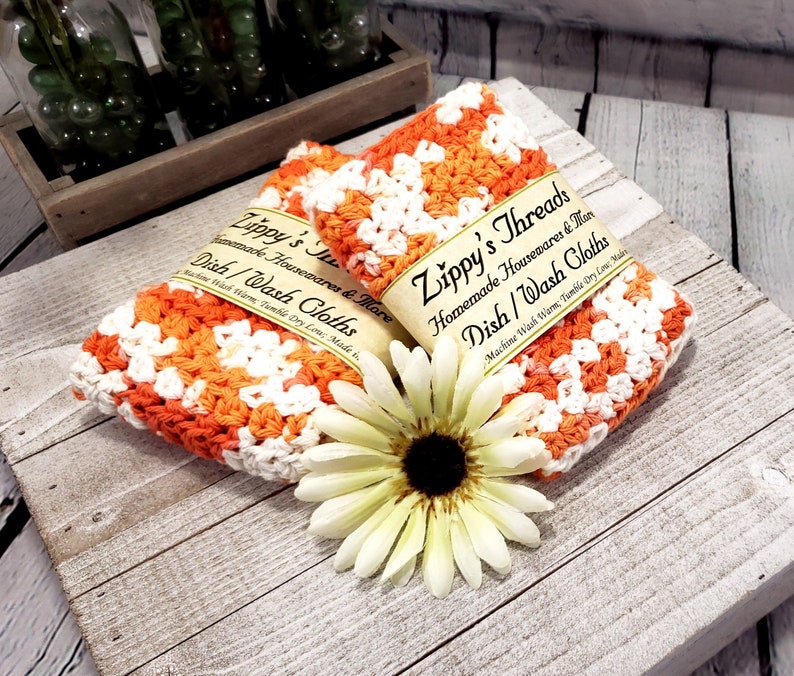 May include: Two handmade dish cloths in orange and white crochet, labeled "Zippy's Threads". The cloths are displayed with a white and yellow flower. The text on the label reads "Homemade Housewares & More Dish/Wash Cloths".