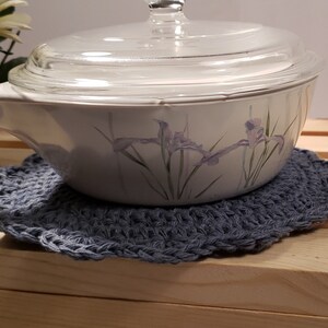 Pode incluir: Uma travessa branca com tampa de vidro, apoiada em um descanso de panela de croch&ecirc; azul feito &agrave; m&atilde;o. A travessa apresenta um desenho floral em roxo e verde. O descanso de panela &eacute; redondo e tem uma borda ondulada.