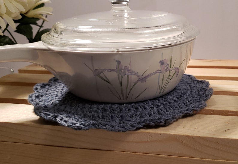 Pode incluir: Uma travessa branca com tampa de vidro, apoiada em um descanso de panela de croch&ecirc; azul feito &agrave; m&atilde;o. A travessa apresenta um desenho floral em roxo e verde. O descanso de panela &eacute; redondo e tem uma borda ondulada.
