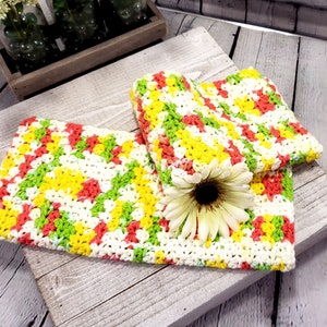 May include: Two crocheted dishcloths in a checkered pattern with white, yellow, green, and pink yarn. The dishcloths are folded and placed on a wooden surface.