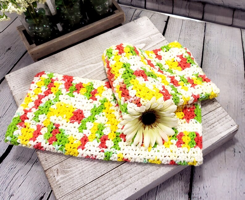 May include: Two crocheted dishcloths in a checkered pattern with white, yellow, green, and pink yarn. The dishcloths are folded and placed on a wooden surface.