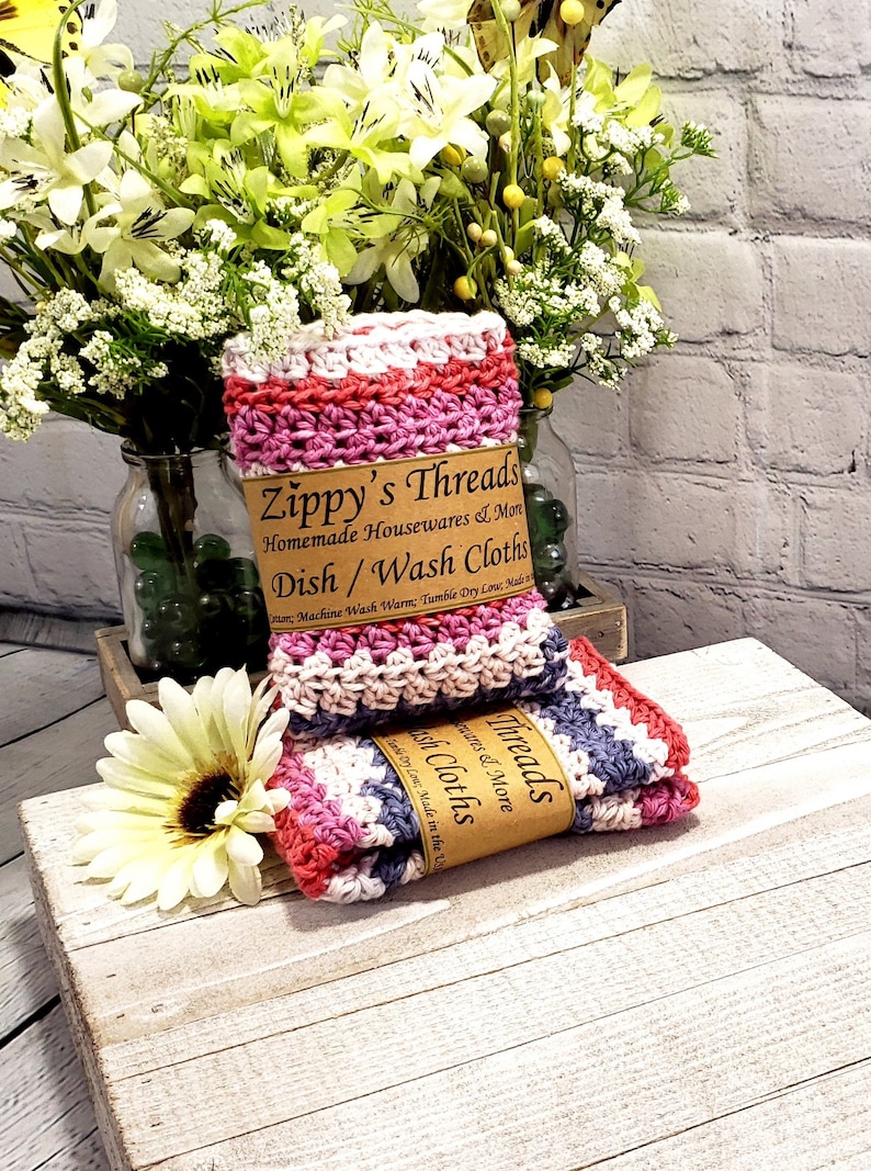 Pu&ograve; includere: Due strofinacci in crochet rosa, bianco e grigio con un'etichetta marrone che recita "Zippy's Threads Homemade Housewares & More Dish/Wash Cloths".