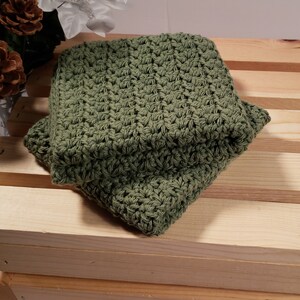 May include: A green crocheted dishcloth or washcloth. The cloth is folded and sitting on a wooden surface.
