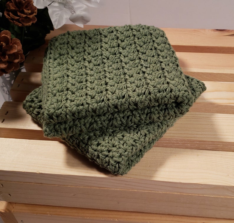 May include: A green crocheted dishcloth or washcloth. The cloth is folded and sitting on a wooden surface.