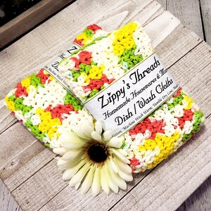 May include: Two crocheted dishcloths in a colourful pattern of yellow, green, pink, and white. The dishcloths are made of cotton and are machine washable. The dishcloths are labelled "Zippy's Threads Homemade Housewares & More Dish/Wash Cloths Cotton; Machine Wash Warm; Tumble Dry Low; Made"