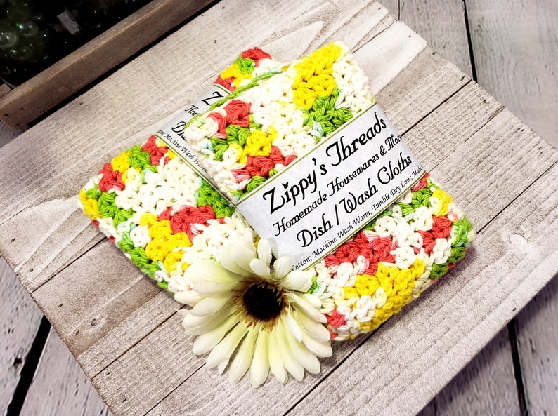 May include: Two crocheted dishcloths in a colourful pattern of yellow, green, pink, and white. The dishcloths are made of cotton and are machine washable. The dishcloths are labelled "Zippy's Threads Homemade Housewares & More Dish/Wash Cloths Cotton; Machine Wash Warm; Tumble Dry Low; Made"