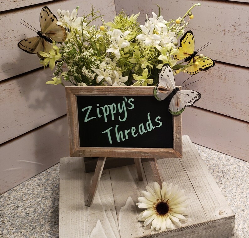 May include: A small chalkboard sign on a wooden easel displays the text "Zippy's Threads" in green script. The sign is surrounded by a floral arrangement with white flowers and decorative butterflies. A white daisy sits in front.