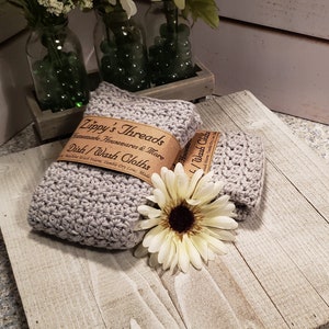 K&ouml;nnte beinhalten: Zwei geh&auml;kelte graue Geschirrt&uuml;cher mit einem braunen Papieretikett mit der Aufschrift "Zippy's Threads Homemade Housewares & More Dish/Wash Cloths". Eine wei&szlig;-gelbe Blume liegt auf einer Holzoberfl&auml;che.