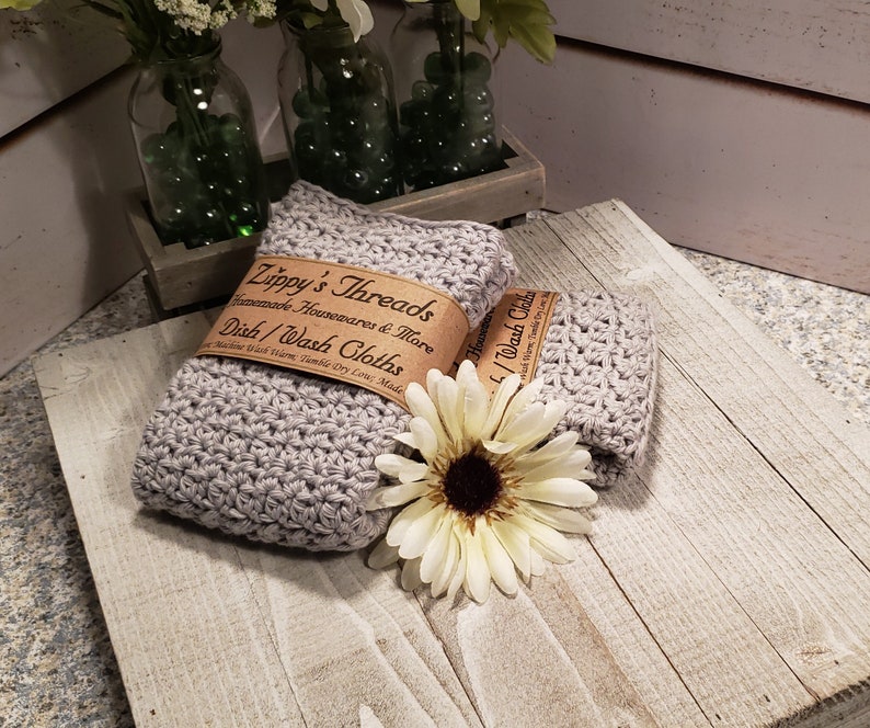 K&ouml;nnte beinhalten: Zwei geh&auml;kelte graue Geschirrt&uuml;cher mit einem braunen Papieretikett mit der Aufschrift "Zippy's Threads Homemade Housewares & More Dish/Wash Cloths". Eine wei&szlig;-gelbe Blume liegt auf einer Holzoberfl&auml;che.